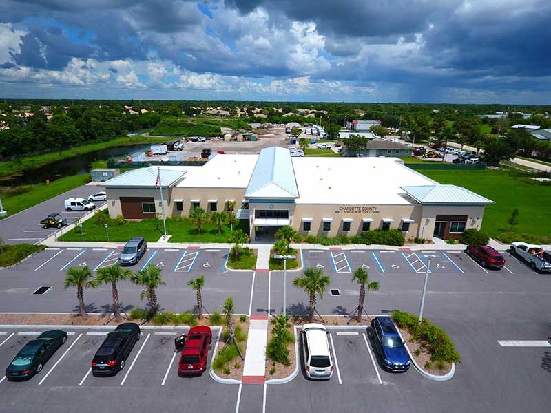 Aerial view of a single-story building with a metal roof in Southwest Florida, surrounded by a parking lot dotted with cars and palm trees under a partly cloudy sky—a showcase of expert Construction Management.