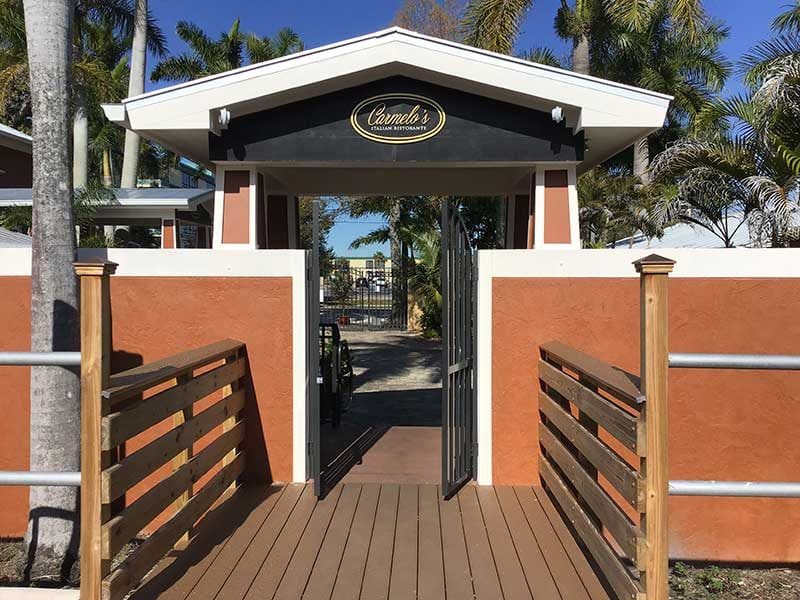 Entrance to a gated outdoor area in Southwest Florida with an overhead sign reading "Camila's," flanked by palm trees and orange walls, and a wooden walkway—showcasing thoughtful construction management—leading up to the gate.