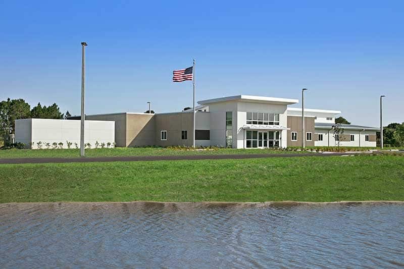 A modern, single-story building with large windows sits behind a grassy lawn and pond in Southwest Florida, with an American flag flying on a pole in front—a testament to expert construction management.