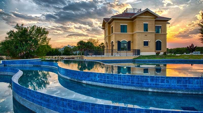 A two-story tan house stands near a curving blue-tiled pool in Southwest Florida, surrounded by trees and a dramatic sunset sky—a perfect blend of beauty and expert construction management.