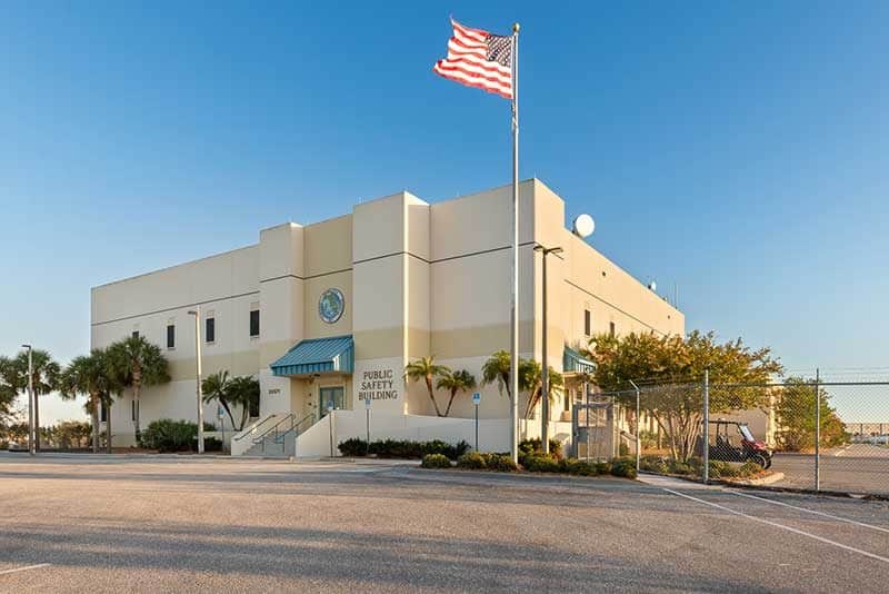 A beige two-story "Public Safety Building" in Southwest Florida stands with an American flag out front, surrounded by palm trees, a parking lot, and a chain-link fence—a testament to expert Construction Management.