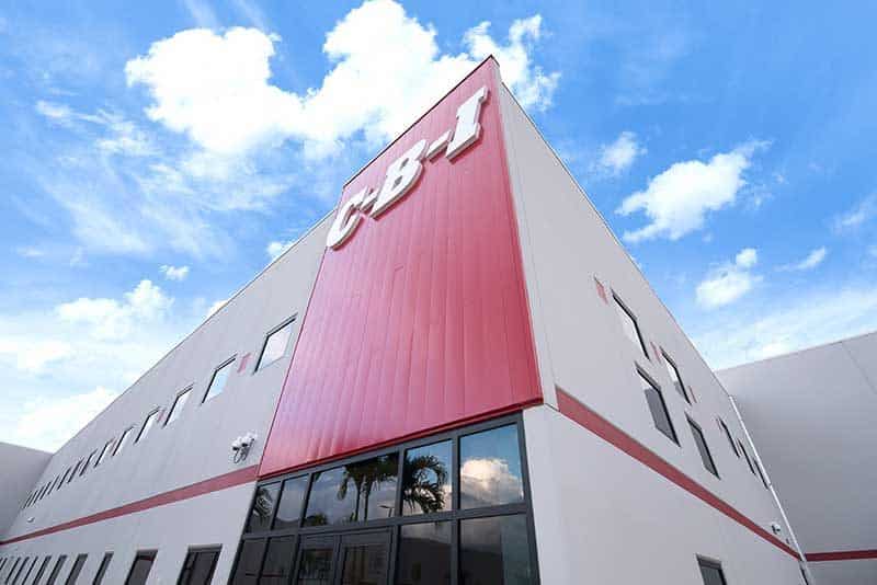 A modern industrial building managed by a leading Construction Management team features a red vertical panel with large white CBI letters, set against a bright blue sky with scattered clouds in Southwest Florida.