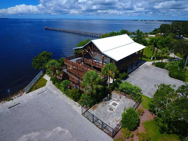 Aerial view of a two-story wooden building with a white roof by a large body of water in Southwest Florida, surrounded by palm trees, greenery, and a nearby pier—an example of expert construction management.
