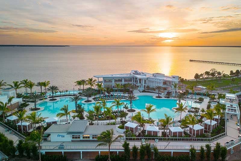 A large waterfront resort in Southwest Florida with palm trees, a pool, and cabanas is shown at sunset, overlooking a calm body of water and a distant pier.