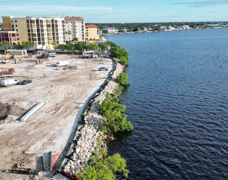 A construction site in Southwest Florida with cleared land borders a body of water, where rocks and small trees line the shoreline; buildings are visible in the background, showcasing expert construction management.