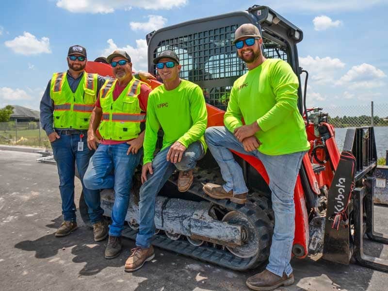 Four construction workers in safety gear and sunglasses stand by a compact track loader on a sunny day at a Southwest Florida work site, exemplifying professional construction management in action.